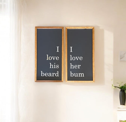 Two framed signs with the text 'I love his beard, she loves his bum' displayed on a wall, with a wooden frame, hanging above a bed.